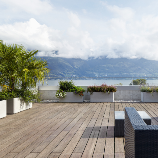 Terrasse en boubou de couleur brune. Un salon de jardin gris foncé est installé, des arbustes verts sont sur la gauche et la vue de la terrasse donne sur un lac avec des montagnes. 