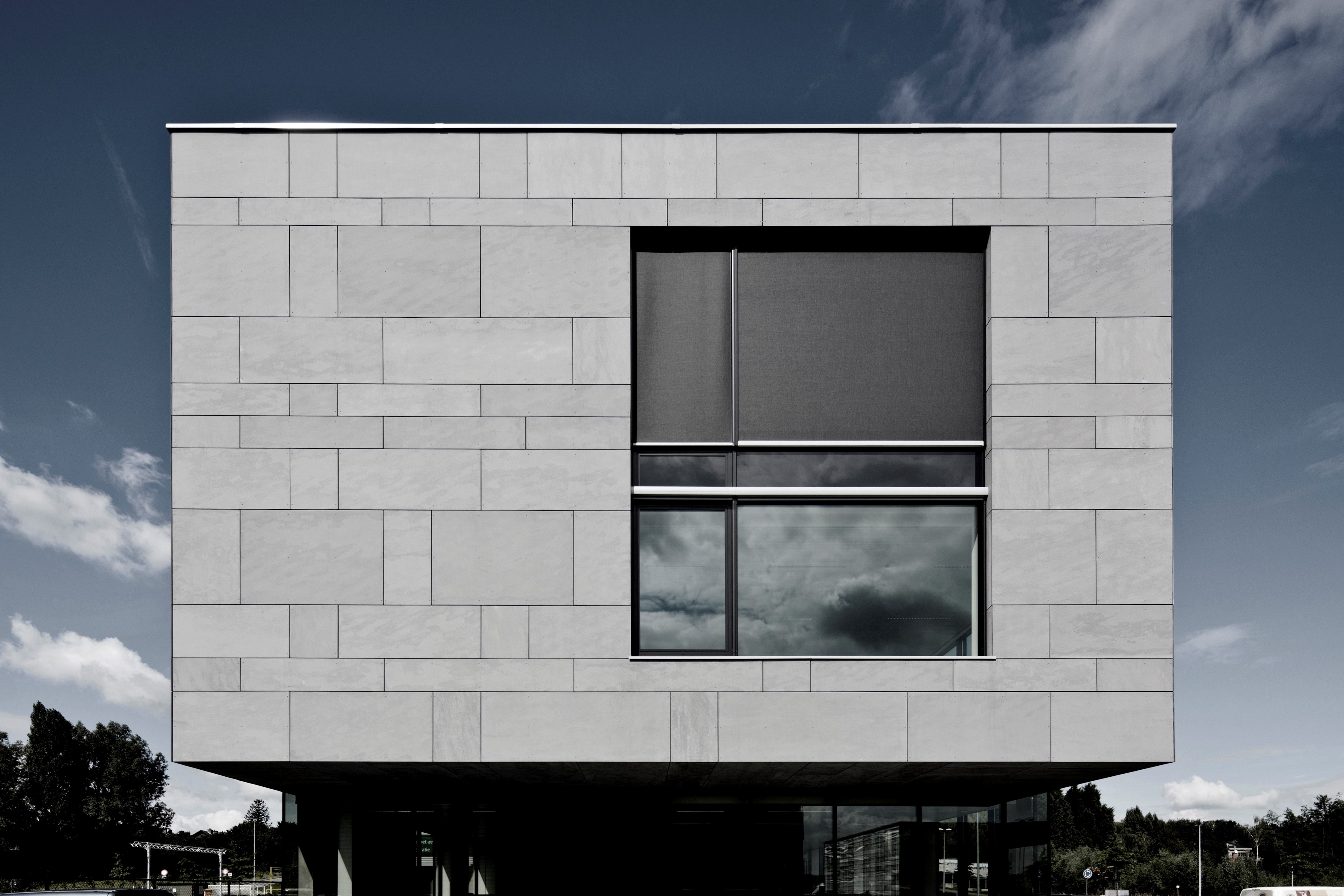 Photo de réalisation. Façade d'un bâtiment avec des panneaux gris et une grande fenêtre sur la droite du bâtiment. 