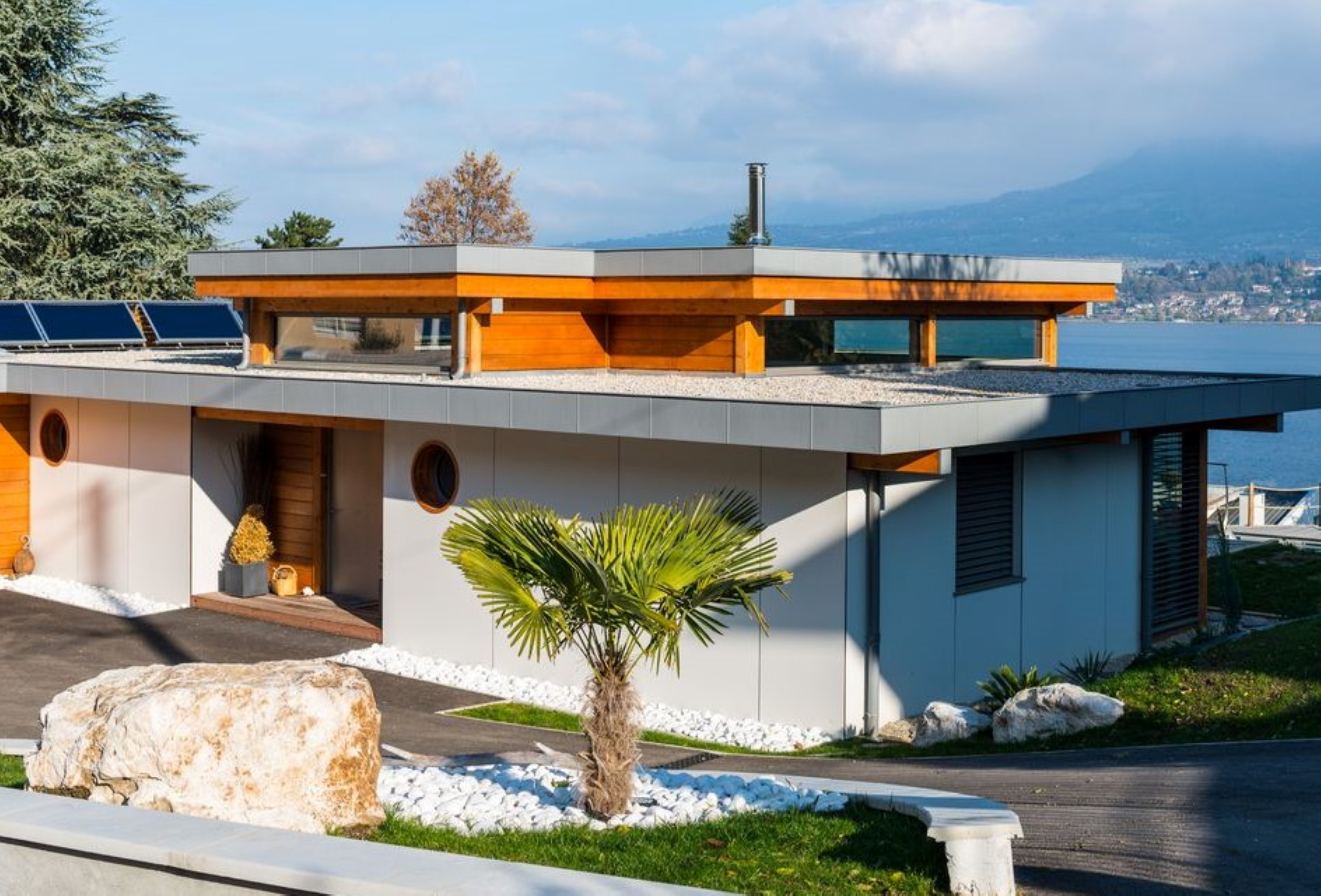 Photo de réalisation. Grande maison toit plat. Panneaux de façade blancs et lames de bardage oranges. Palmier devant la maison et la mer en arrière-plan.